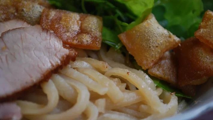 Close-up of cao lau: thick noodles, slices of roast pork, crisp fried pieces and fresh greens